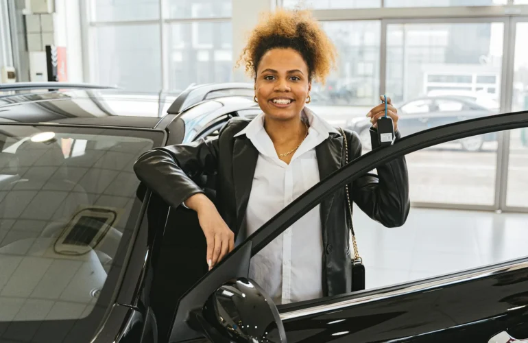 Smiling woman with car keys standing beside her new vehicle in a dealership showroom.